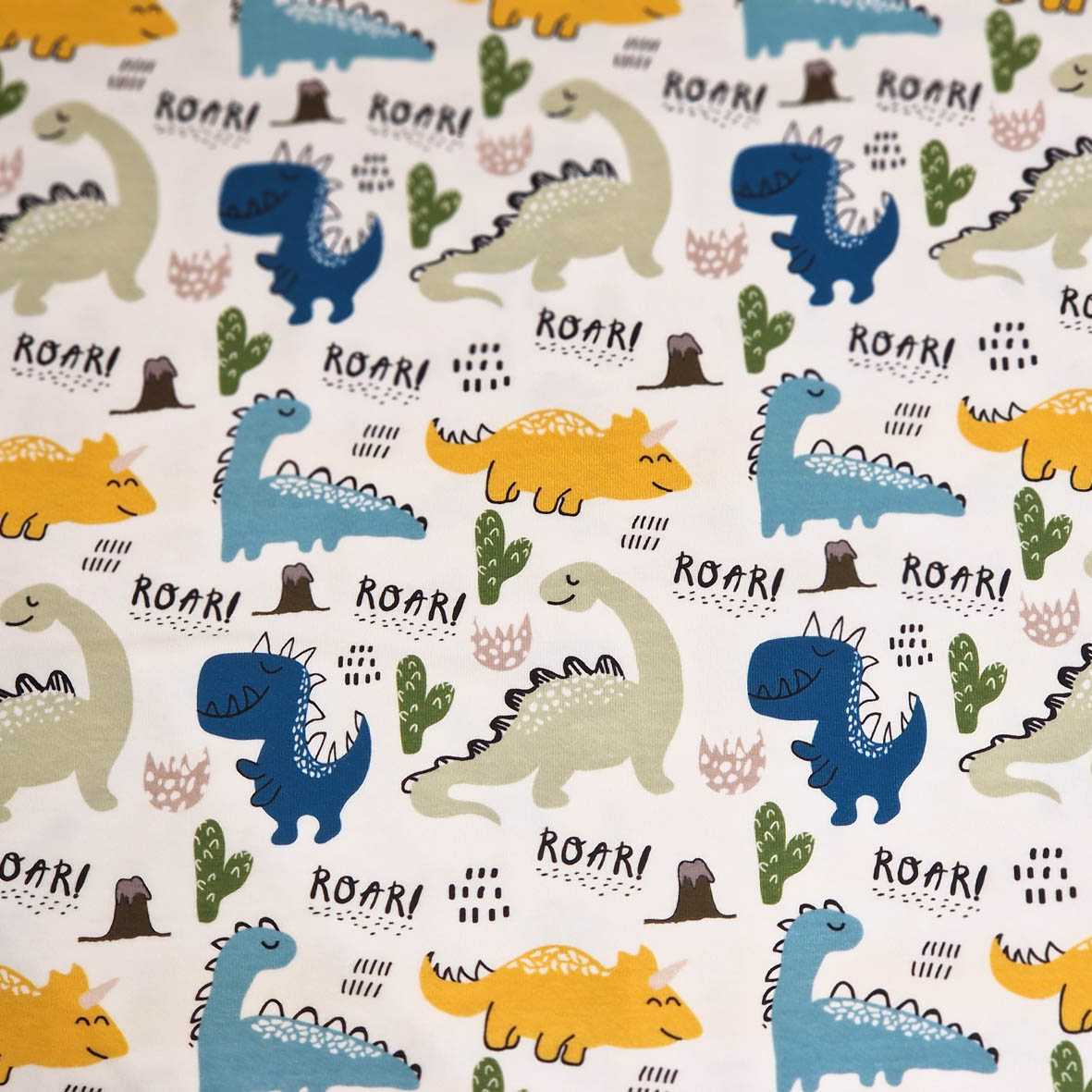 Stoff von bunten Dinosauriern und Kakteen auf weißem Hintergrund mit dem Schriftzug "ROAR!" in verschiedenen Farben.