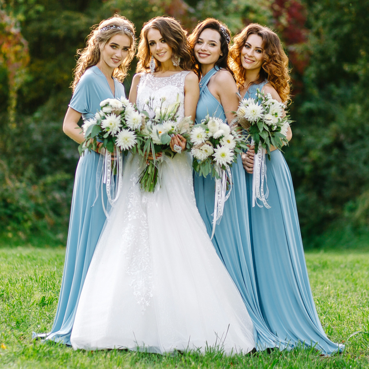 Vier Frauen in blauen Kleidern und einer Braut in weißem Kleid, umgeben von grüner Wiese und Blumensträußen.